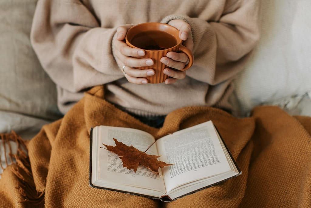 Skip the Sales and Focus on You This Weekend: It’s OK 1 A woman enjoys a cozy autumn day, holding a tea mug over an open book with a maple leaf. skip the sales